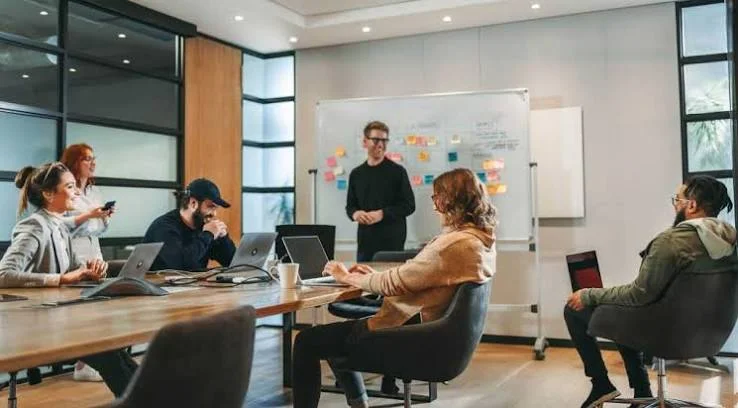 A team working session around a conference table, with a facilitator at a whiteboard of sticky notes